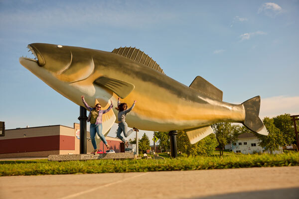 Two women jumping in the air in front of Baudette's Willie the Walleye statue