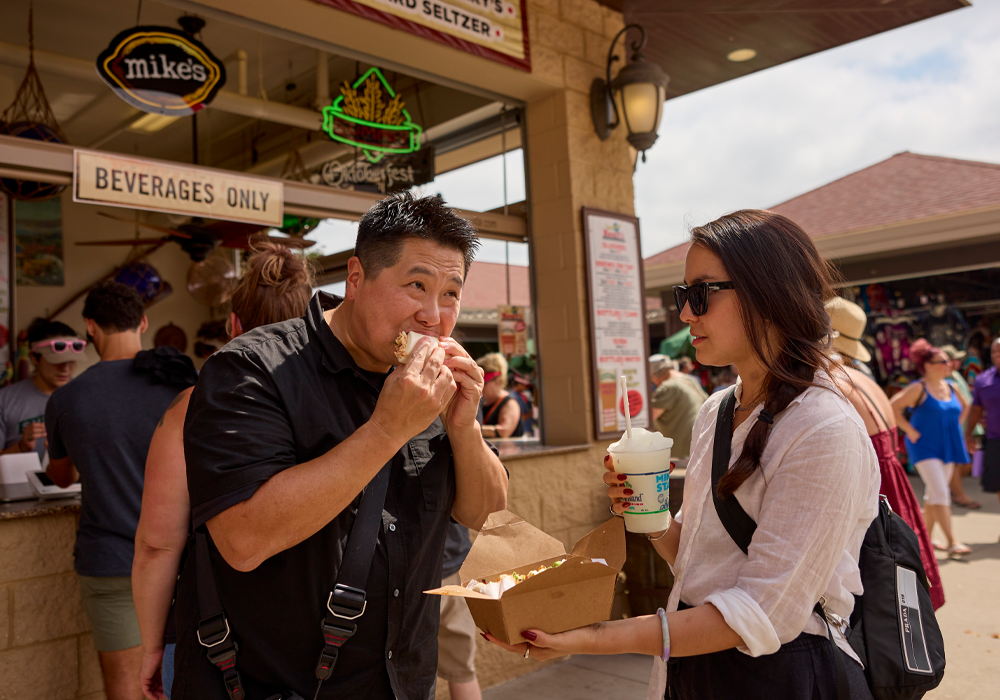 two friends at the State Fair. One is holding a Mike's Hard Lemonade Slushie and a paper to-go box, the other is biting into a bao bun.