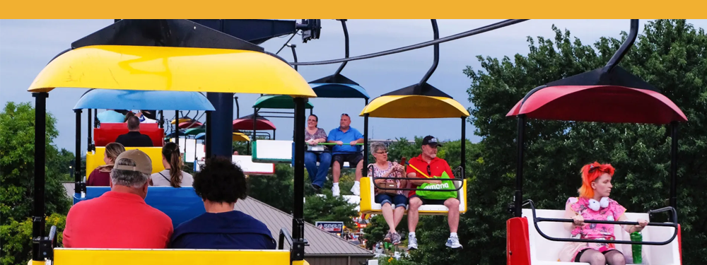 POV from someone on the SkyGlider – visual of people riding both directions in yellow, red, and blue cars. green trees are at riders' eye level.
