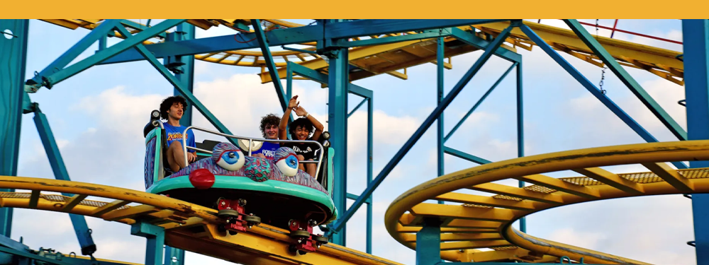 a group of three younger people in a crazy mouse cart, raising their hands up while they ride the yellow and teal rollercoaster track.