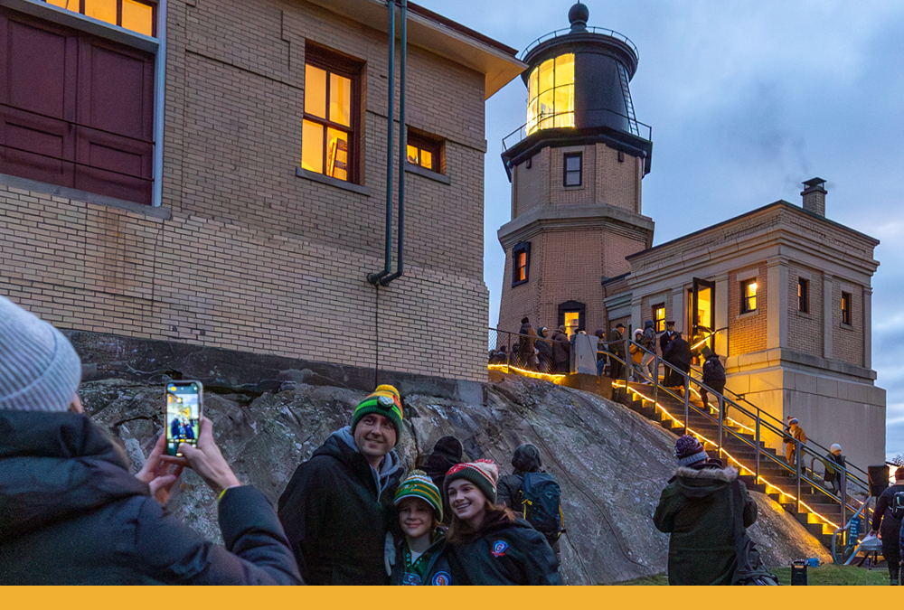A family takes a photo at dusk with the beacon of Split Rock Lighthouse lit. People walk up the illuminated stairs behind them.