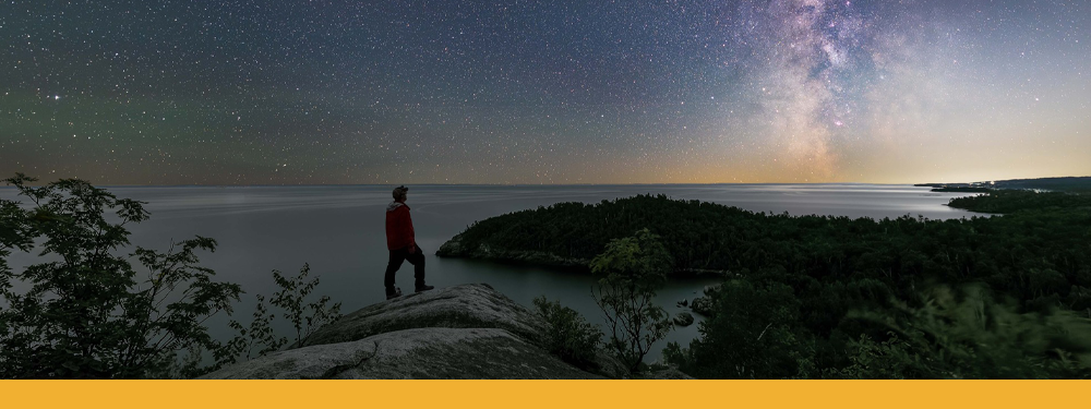 Hayes' silhouette on a rock cliff. A beautiful blue, purple, pink, and orange sky covered in stars is on the horizon above Lake Superior.