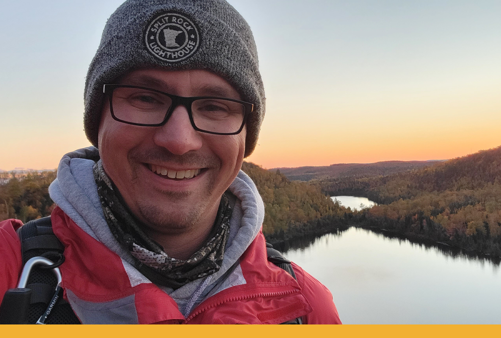 Hayes, wearing a knit Split Rock Lighthouse hat and warm red jacket, smiles in a selfie while hiking. Two lakes can be seen behind him.