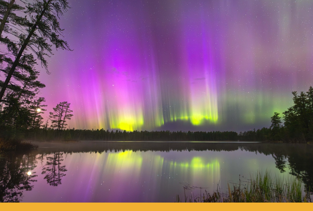 Vibrant purple, pink, and neon green aurora lights in the sky are also reflected in a lake below. The ribbons of color dance across the sky.