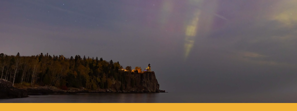 Split Rock Lighthouse on the cliff. The calm grey water blends into the the night sky. Faint yellows and purples of the aurora streak down.