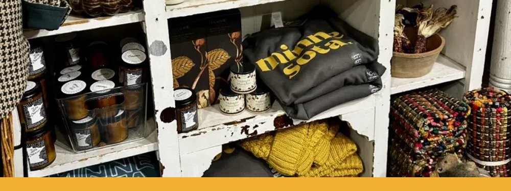 white shelves filled with candles, yellow knit hats, and grey shirts with a fat yellow "minnesota" serif font