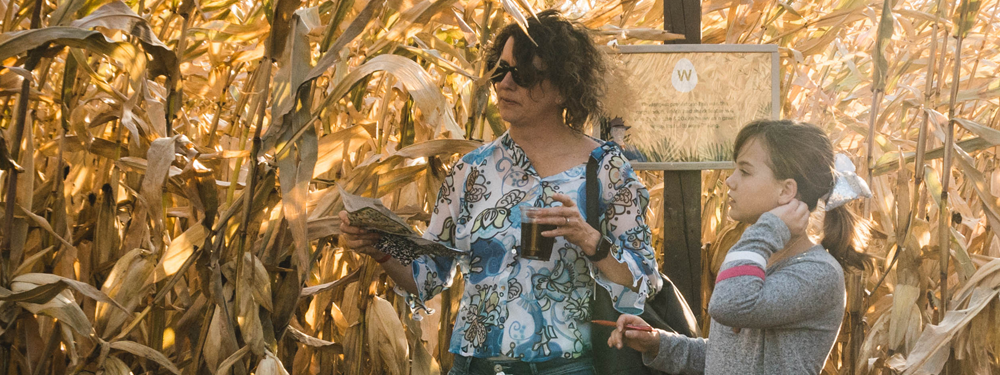 a mother holds a map and a drink while her daughter holds a pencil. Corn stalks blow behind them with a sign behind them.