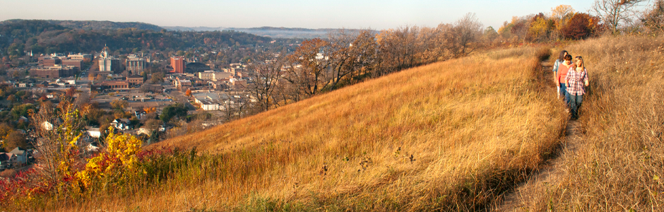 3 people hiking on a grassy bluff golden with sunlight. Below is the quaint town of Red Wing.