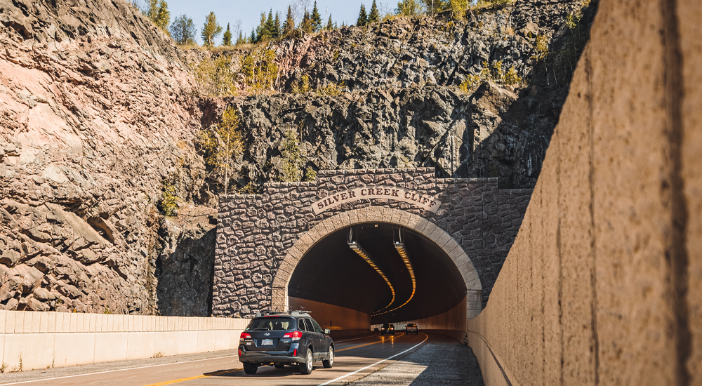 Cars driving through an arched tunnel made through a dusty red and grey cliff on the North Shore. Green pines grow on top of the cliff above.