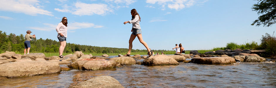 several people rock hopping over the shallow headwaters on a sunny day