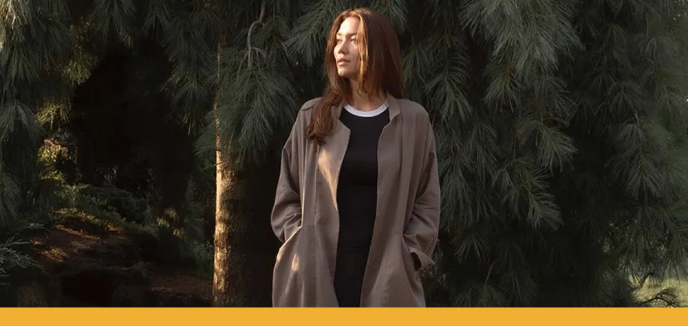 woman standing in front pine trees wearing a warm brown hand-sewn coat