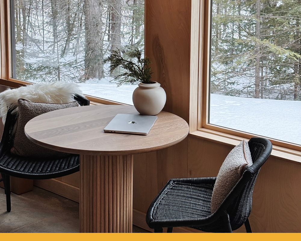 a wooden round table with two black wicker chairs next to two windows inside a cozy cabin. Outside snow has powdered the ground.