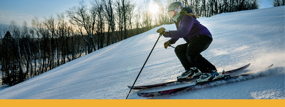 woman downhill skiing in a green helmet, rainbow goggles, purple winter coat, black snow pants, and yellow gloves.