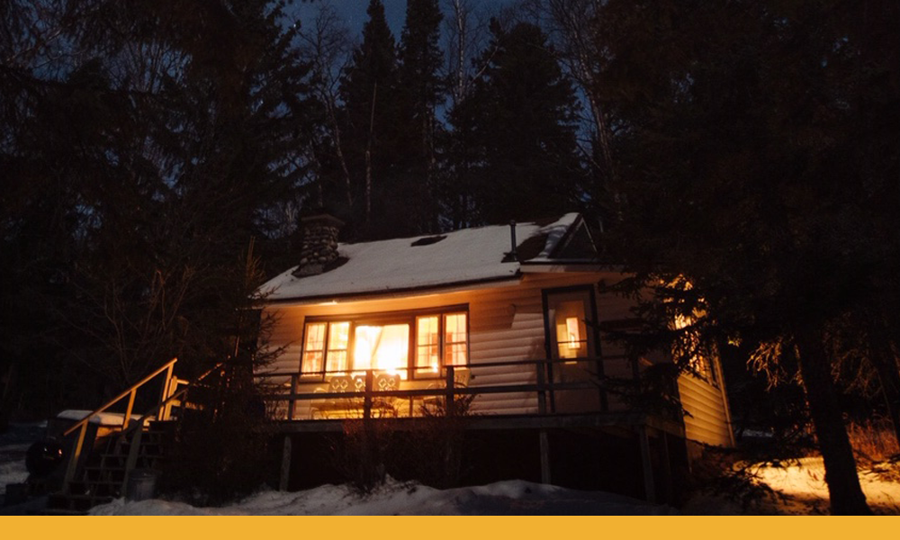 small cottage's warmly lit interior glows outside amongst the night snow and tall pines.