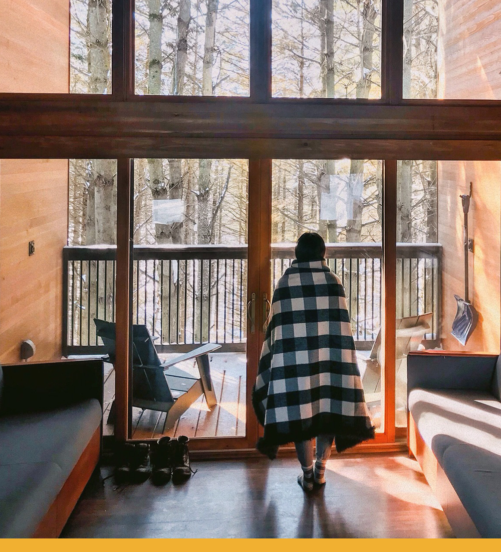 person wrapped in a black+white plaid blanket & warm socks looks out an all window side of a cabin. The sun casts dreamy shadows on the interior.