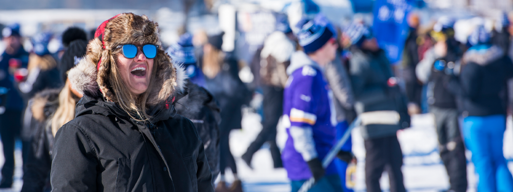 laughing person in large black winter coat, red plaid bomber hat, and reflective sunglasses is on the ice with a large crowd