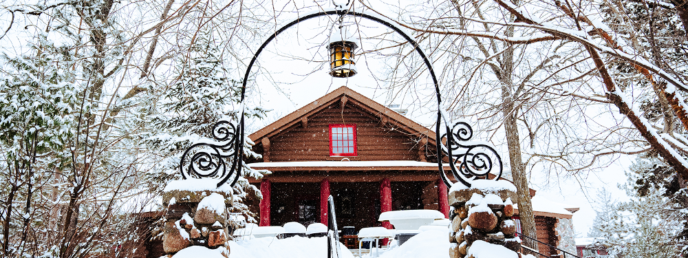 Iron archway with a hanging lantern lit in front of a log cabin with red shutters. Snow lightly falls on already covered ground.