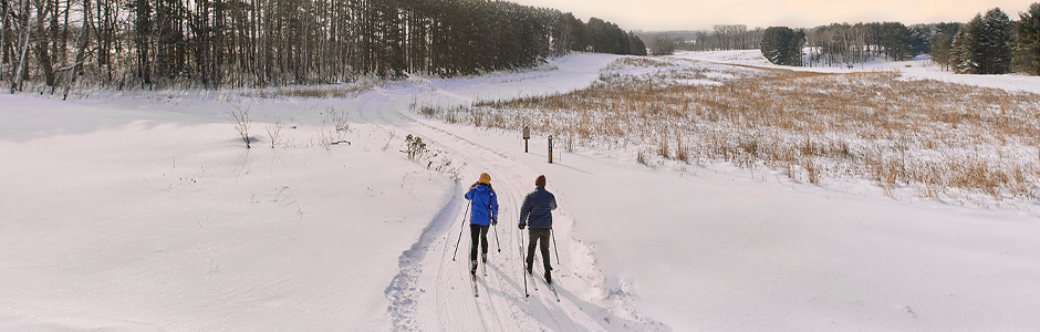 the backs of two people in winter gear, glide on cross-country skis on white powdery snow. The sun rising in the background.