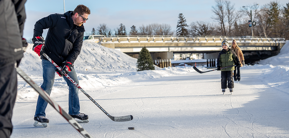 Two adults and two kids in winter coats, gloves, and hockey skates, play a friendly game of hockey on an outdoor ice rink.