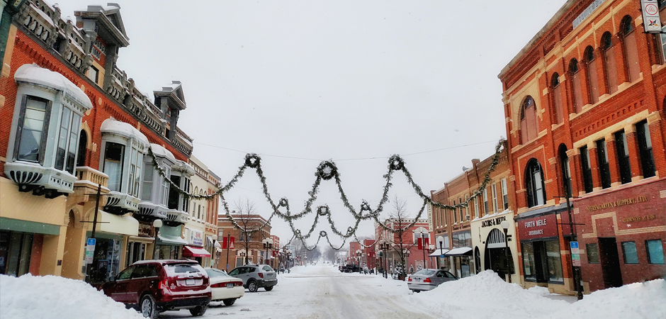a small town main street looks like a postcard covered in fresh snow, with a garland of wreaths strung from one side of the street to the other.