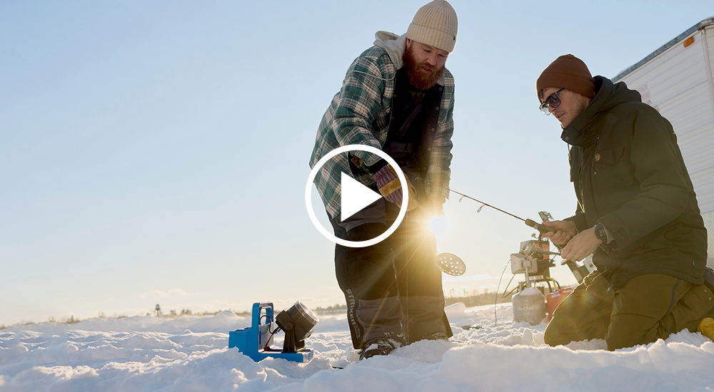 Two men outside on big open lake frozen over, peak into a hole in the ice, their fishing poles hovering in their hands.