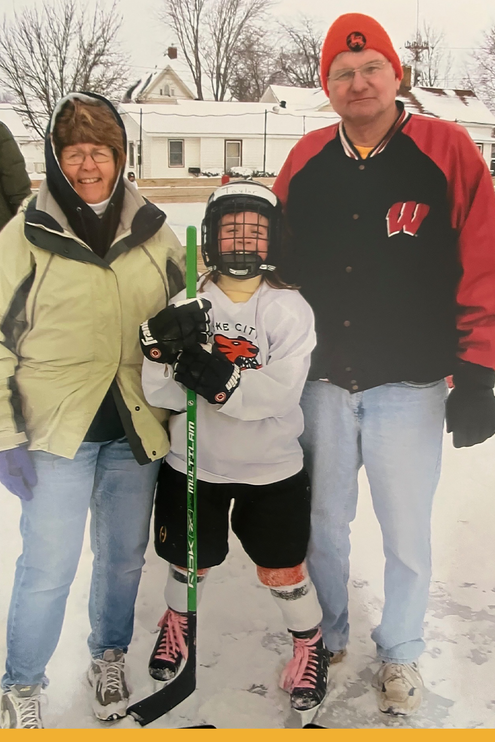Taylor at a young kid smiling with full hockey gear on while holding her stick on an outdoor ice rink.