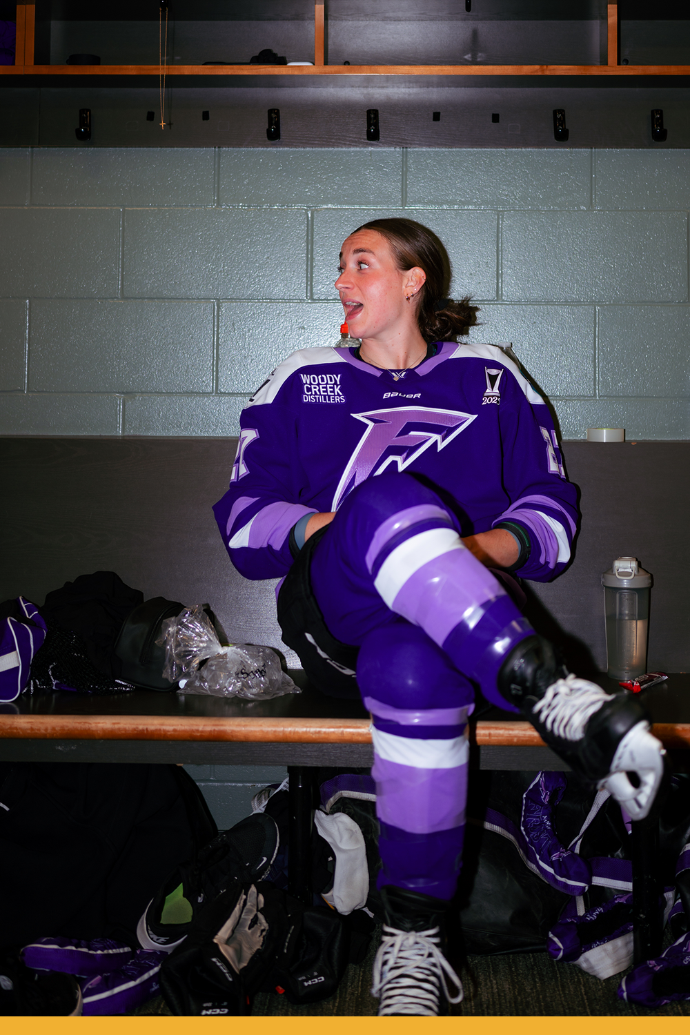 Taylor talking to someone off camera in the locker room. She's sitting on the bench with her skates crossed. Pads, water, and ice can be seen.
