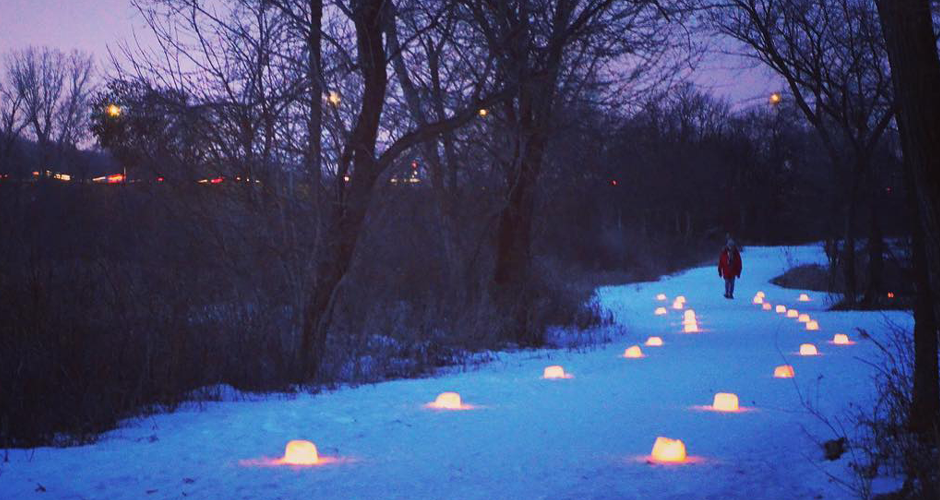 person in winter gear walking down a snow trail designated by spherical ice luminaries along the sides. a purple night sky casts shadows on trees.