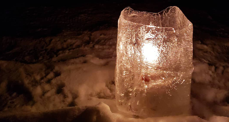 a candle's warm orange light refracts through a cylindrical ice wall from a luminary sitting in a snow bank.