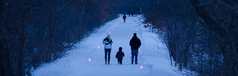 Two adults with a bundled up toddler walk at dusk into the woods following along a snowy, candlelit trail.