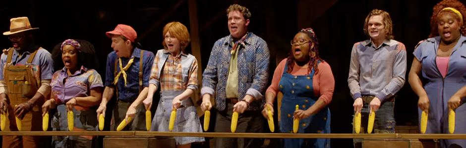 a chorus line of actors dressed in farmer's overalls and plaid, hold corn on the cob in both hands.