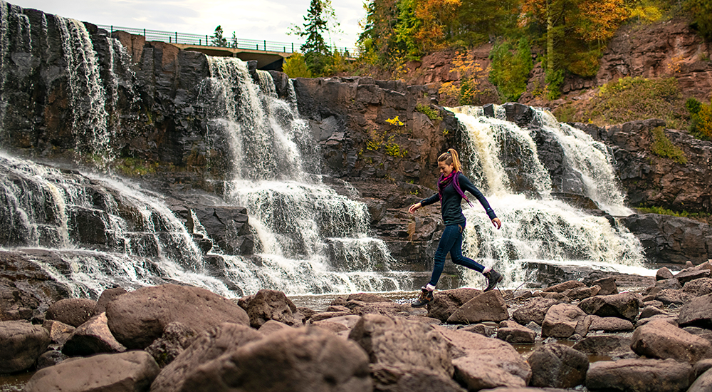 A woman delicately hops from one rock to another, a wide waterfall streaming over rocks is in the background with fall trees.