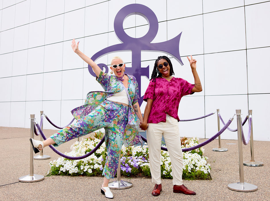 two people dramatically pose with cheesy smiles in front of a purple statue of Prince's name from 1993-2000. 