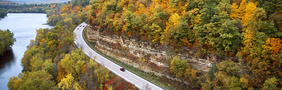 a car on a winding road around a bluff. blue water is to the left of the road, fall trees and a red bluff are to the right.