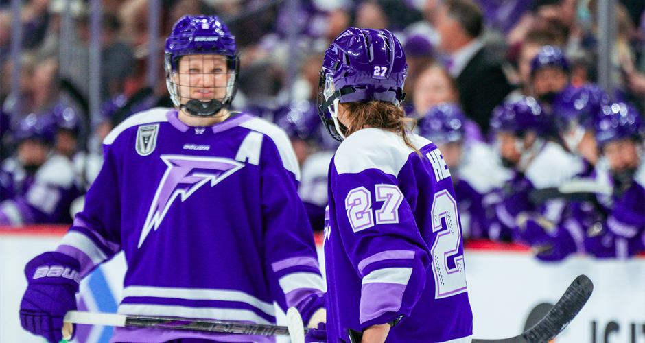 two professional female hockey players in purple quickly communicate on ice before the puck drops