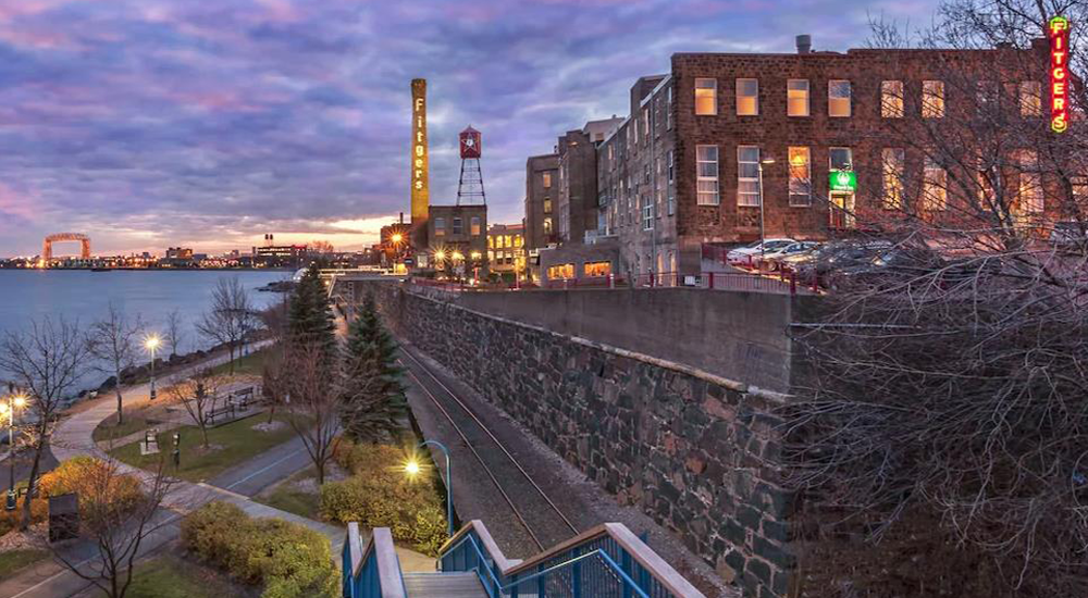 View of Fitger's Brewing brown brick building. A purple and pink cloudy sky and dusk and Duluth's lift bridge in the background.