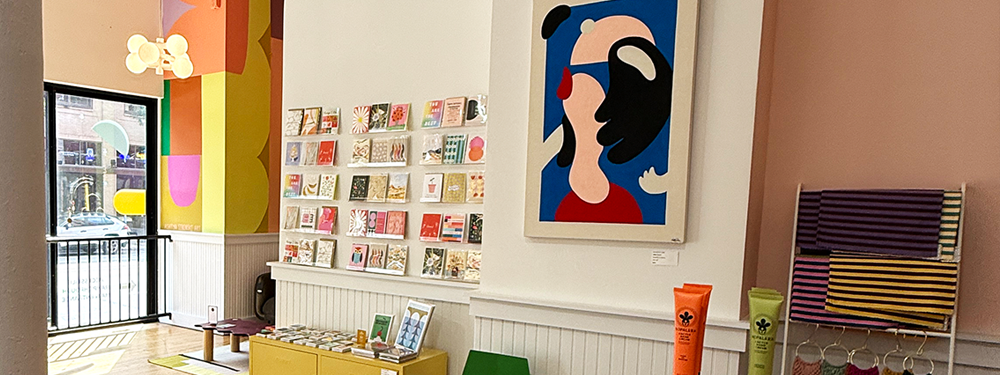 Warm store interior featuring a wall of handmade cards, large scale paintings, and other giftables on shelves.