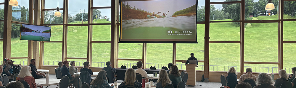 a gathering sits around round tables while looking at a presentation and speaker. behind the speaker, giant windows show well kept green fields.