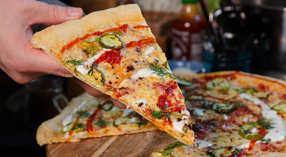 a hand holding one triangle slice of homemade pizza with pickles, peppers, dill Havarti, ham, sriracha and ranch.