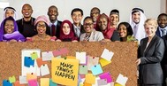 Photo of a diverse group of people standing by a large bulletin board that has a note saying Make Things Happen on it