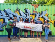 Masked people holding a Spark-Y banner in front of a mural