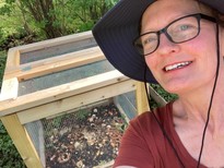 A woman in a wide-brimmed hat taking a selfie in front of a backyard compost bin