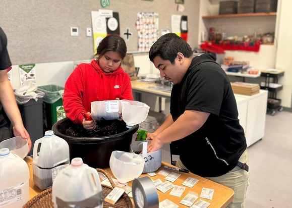 A young man and woman in a classroom putting dirt from a large black bucket into cut open gallon milk jugs