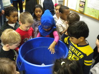 A group of preschool aged kids gathered around a recycling cart