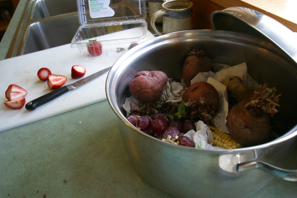 Bowl on counter with potatoes, grapes, corn, and other food scraps next to cutting board with strawberries
