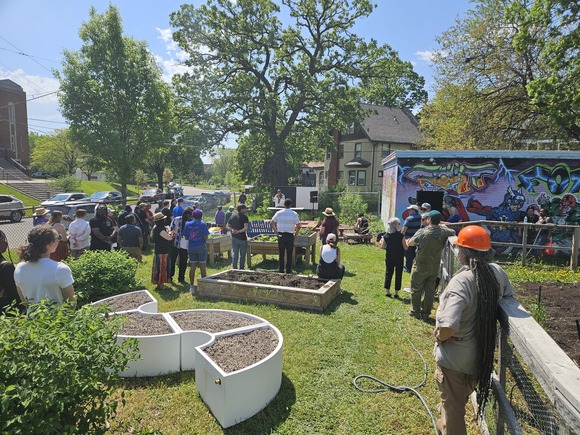 Group of people gathered in a small park with raised garden beds facing a stage