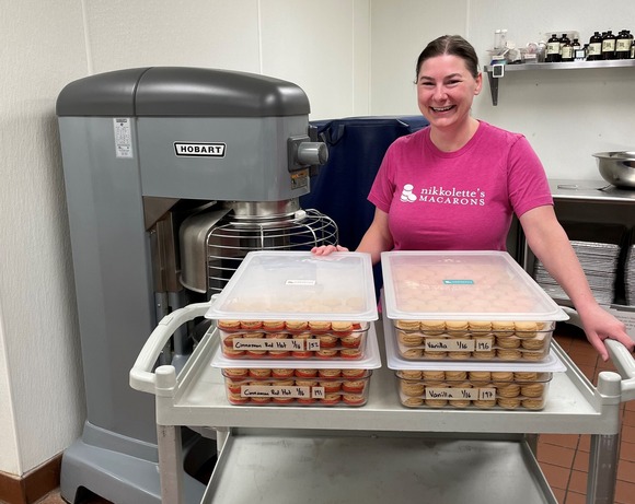 Nikkolette's Macarons owner standing with reusable bakery boxes filled with macarons on a cart in commercial kitchen