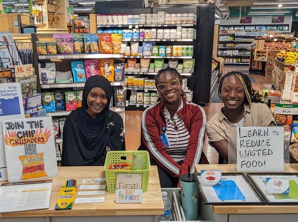 Three Green Pathways interns sitting at a table at a food coop with signs about reducing food waste and promoting the Stop Food Waste Challenge