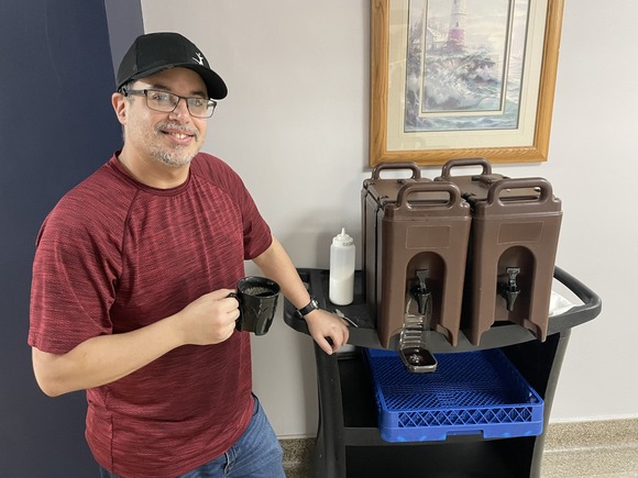 Man holding a reusable coffee cup next to a cart with two large carafes of coffee and a try of reusable coffee mugs