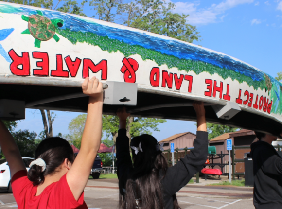 Youth with MGIZI holding canoe over their heads painted with water and turtle and words protect the land and water